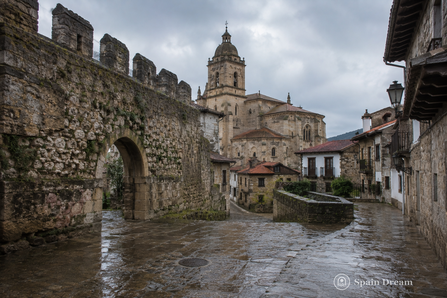 Средневековая стена, базилика Santa María del Conceyu и каменные улицы старого Льянеса после дождя